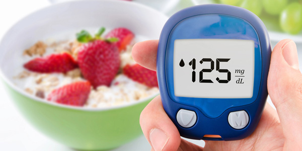Hand hält ein blaues Blutzuckermessgerät mit Anzeige 125 mg/dL. Im Hintergrund steht eine grüne Schüssel mit Müsli und Erdbeeren. Darstellung kann auf Diabetesberatung und Ernährungskontrolle hinweisen.
